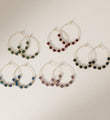 Five pairs of 30mm gold plated hoop earrings with faceted glass beads in black, green, burgundy, blue and purple on a neutral background