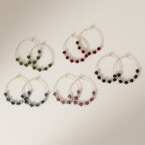 Five pairs of 30mm gold plated hoop earrings with faceted glass beads in black, green, burgundy, blue and purple on a neutral background
