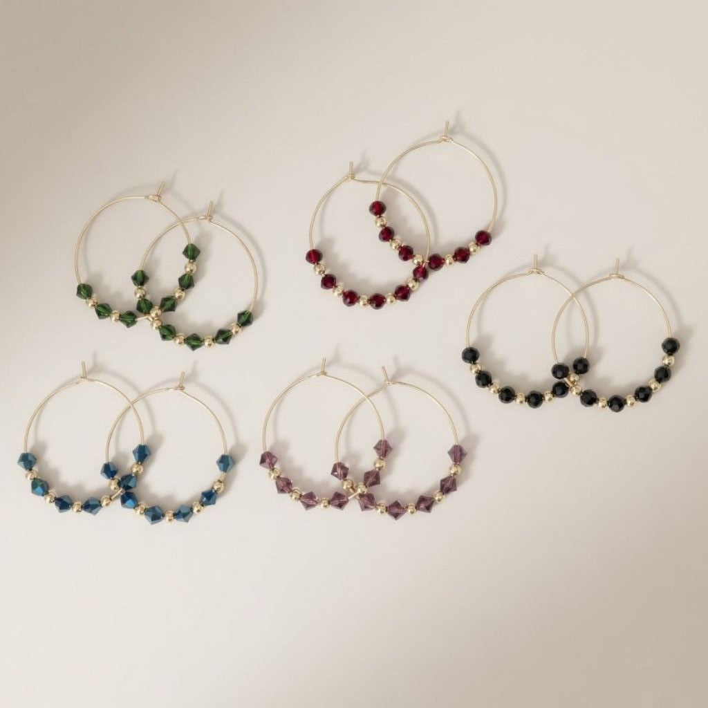 Five pairs of 30mm gold plated hoop earrings with faceted glass beads in black, green, burgundy, blue and purple on a neutral background