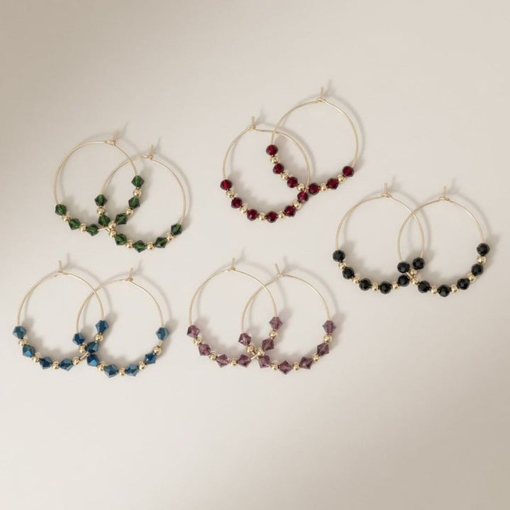 Five pairs of 30mm gold plated hoop earrings with faceted glass beads in black, green, burgundy, blue and purple on a neutral background