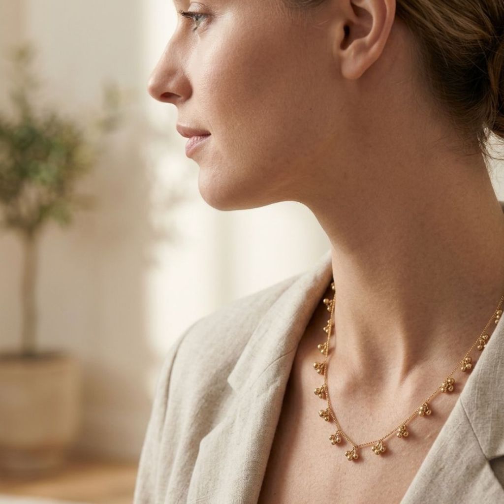 Side profile of a woman wearing a gold ball cluster charm necklace with a linen blazer in a bright interior