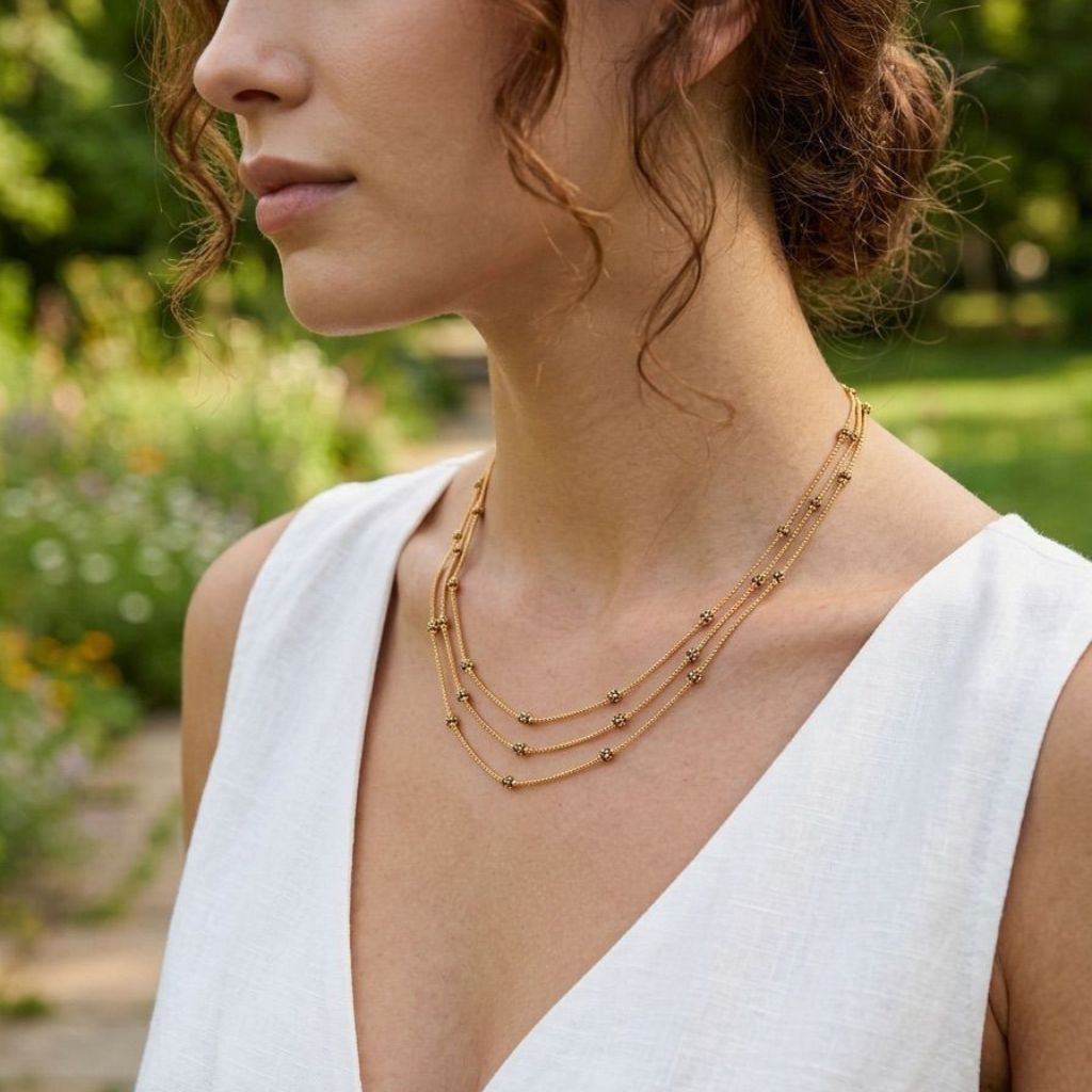 Side profile of a woman wearing an 18ct gold plated triple strand black and gold bead necklace with a white sleeveless top outdoors